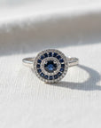 Anillo Sophia Schneider Joyas de oro blanco 18K con zafiro central y doble halo de zafiros y diamantes brillantes.