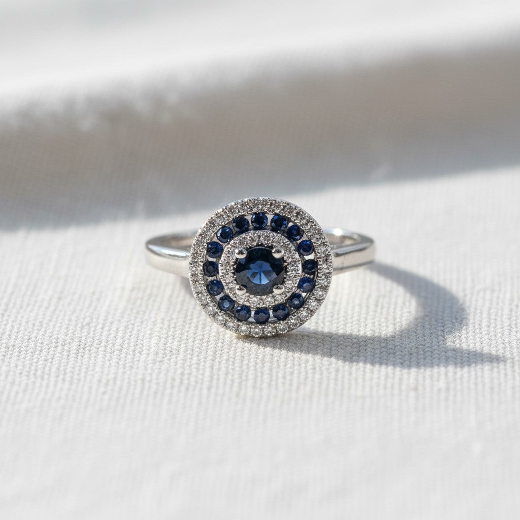 Anillo Sophia Schneider Joyas de oro blanco 18K con zafiro central y doble halo de zafiros y diamantes brillantes.