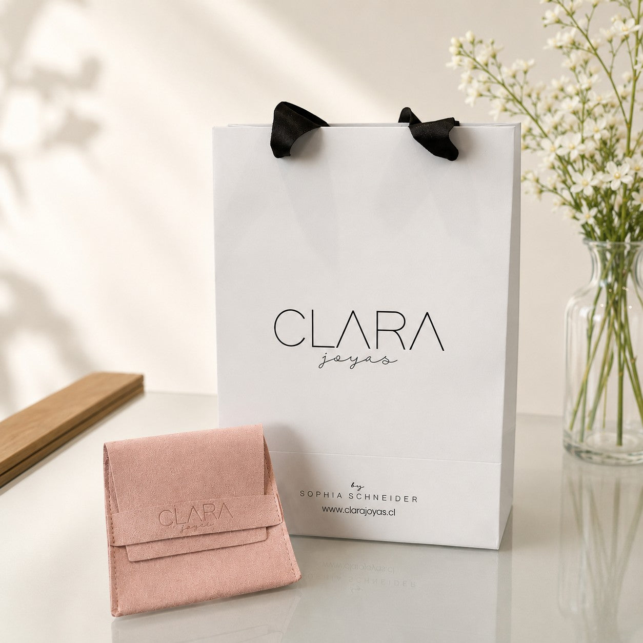 Bolsa de regalo blanca y sobre de gamuza rosa de Sophia Schneider Joyas sobre una mesa con flores de fondo