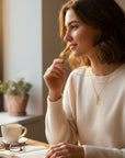 Mujer con suéter beige usando un colgante de cruz de oro 18K de Sophia Schneider Joyas junto a una ventana